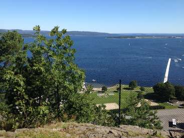 A scenic view of a deep blue lake, with lush green trees in the foreground, grassy park area, distant islands, and clear skies above. Peaceful and inviting.