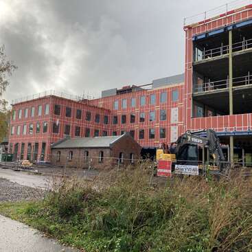 A large modern building under construction with red exterior panels surrounds a small older brick structure. Construction equipment and materials are visible; cloudy sky overhead.