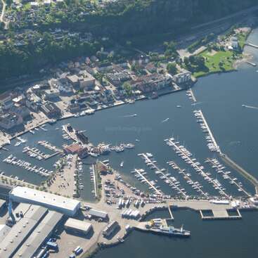 The image depicts an aerial view of a marina, featuring numerous boats and buildings, with a body of water and a town in the background, surrounded by trees.