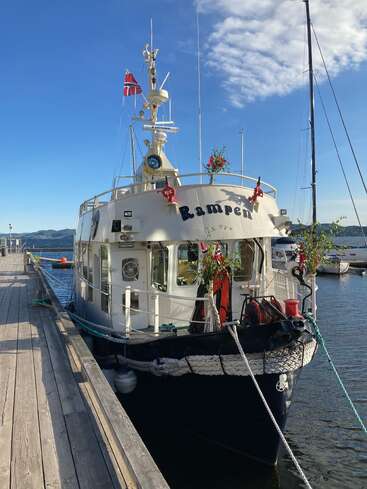 Ein weißes Boot namens „Rampen“ liegt an einem hölzernen Steg, der mit Blumen und norwegischen Flaggen geschmückt ist, unter einem strahlend blauen Himmel mit leichten Wolken.
