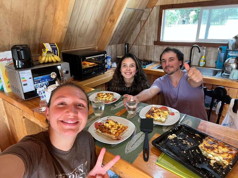 Trois personnes sont joyeusement réunies autour d'une table de cuisine en bois ; elles dégustent des parts de pizza ou de quiche maison, sourient et prennent la pose pour un selfie de groupe plein de bonne humeur.