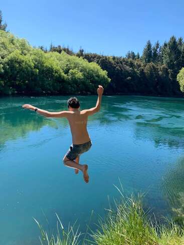 A boy in swim trunks jumps enthusiastically into a clear blue river, surrounded by lush green trees and grass under a bright, sunny sky. Summer fun!