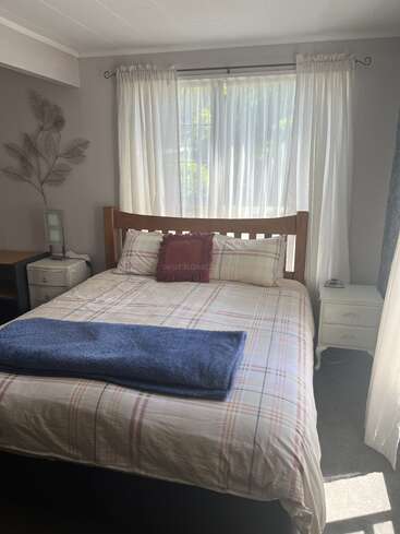 A cozy bedroom with a plaid bedspread, wooden headboard, blue blanket, white curtains letting sunlight in, side tables, and decorative wall art with leaf motifs.