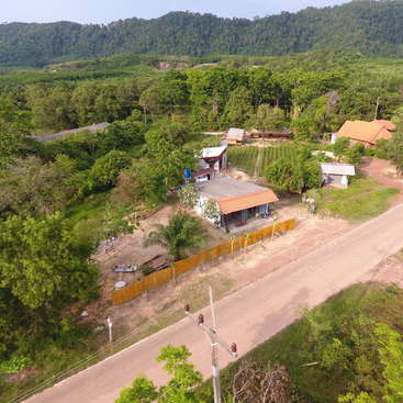 La imagen representa un paisaje rural con una carretera, casas y árboles, sobre un fondo de exuberante vegetación y un terreno montañoso bajo un cielo azul despejado.