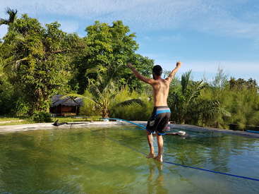 La imagen muestra a un hombre caminando en una cuerda de equilibrio sobre un estanque, con los brazos en alto, rodeado de exuberante vegetación y un sereno cielo azul.
