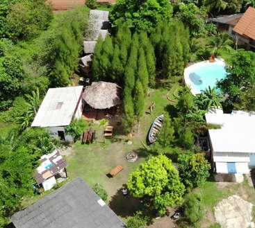 La imagen representa un entorno tropical sereno, con una piscina, estructuras de techo de paja, árboles frondosos y un barco, rodeado de un ambiente tranquilo y una vegetación vibrante.