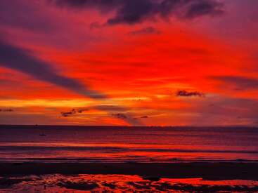La imagen representa una serena puesta de sol sobre el océano, con vibrantes tonos rojos y naranjas que iluminan el cielo y se reflejan en la tranquila superficie del agua.