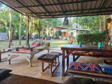 La imagen muestra un sereno patio exterior con una mesa y bancos rústicos de madera, rodeado de exuberante vegetación y una casa al fondo.
