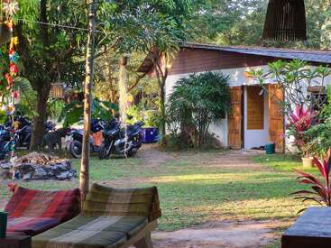 La imagen representa un entorno sereno al aire libre con un edificio blanco, árboles frondosos y plantas vibrantes, con algunas motos aparcadas al fondo.