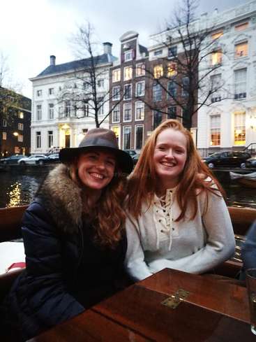 A imagem mostra duas mulheres sentadas em um barco em um canal, com uma paisagem urbana com prédios e carros ao fundo, em um céu nublado.
