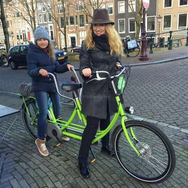 A imagem mostra uma mulher e uma criança ao lado de uma bicicleta verde-limão em uma rua de paralelepípedos, com prédios ao fundo.