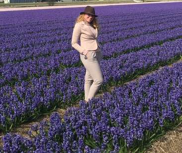 A imagem mostra uma mulher em pé em um campo de flores roxas, vestindo um terno bege e chapéu marrom, com as mãos nos bolsos e olhando para baixo.