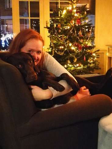 A imagem mostra uma mulher sentada em um sofá com um cachorro marrom, em frente a uma árvore de Natal, com uma janela e uma porta atrás deles, tudo em um ambiente aconchegante de sala de estar.