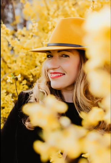 A imagem mostra uma mulher com longos cabelos loiros, usando um chapéu amarelo e uma blusa preta, em frente a um arbusto ou árvore amarelo vibrante, com flores amarelas borradas em primeiro plano.