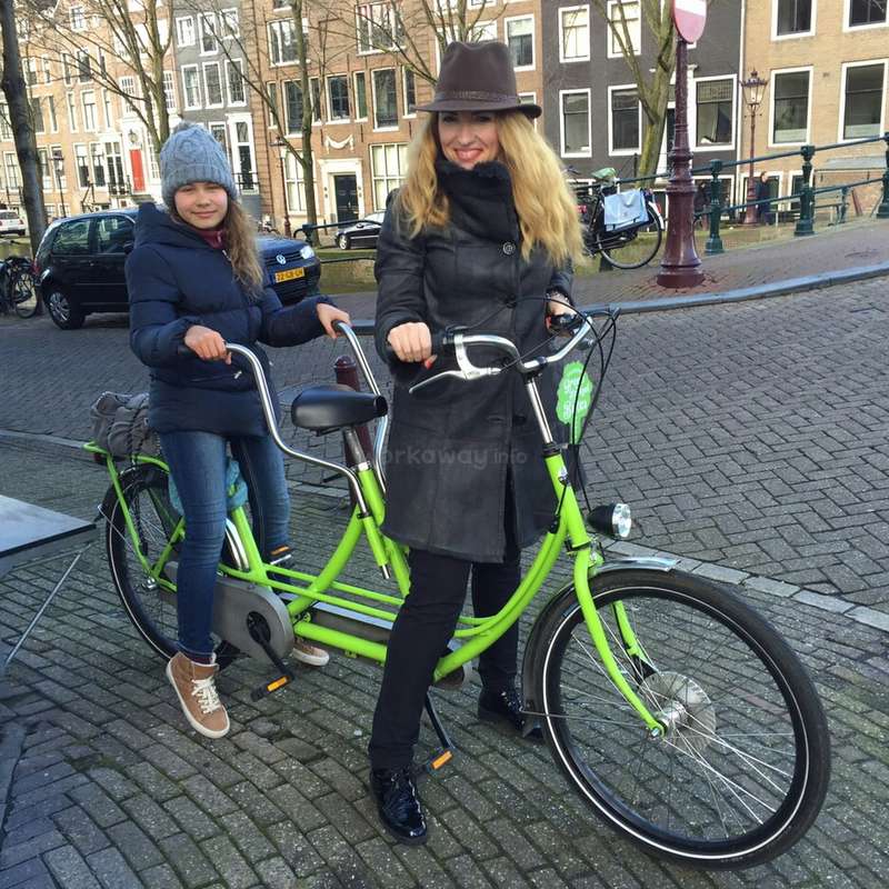 A imagem mostra uma mulher e uma criança ao lado de uma bicicleta verde-limão em uma rua de paralelepípedos, com prédios ao fundo.