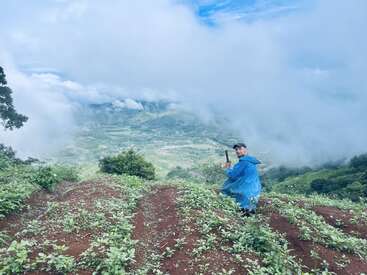 The image depicts a man in a blue raincoat and black hat, kneeling in a field of green plants, holding a stick, with a mountainous landscape and cloudy sky in the background.