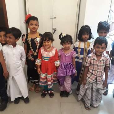 The image shows a group of children standing in a line, dressed in various colorful outfits, posing for a photo in front of a white wall with a door.