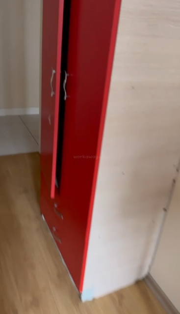 Une armoire rouge à deux portes, dotée de poignées argentées, trône sur un sol en bois, près d'un mur blanc. La pièce semble bien rangée et baignée d'une douce lumière naturelle.