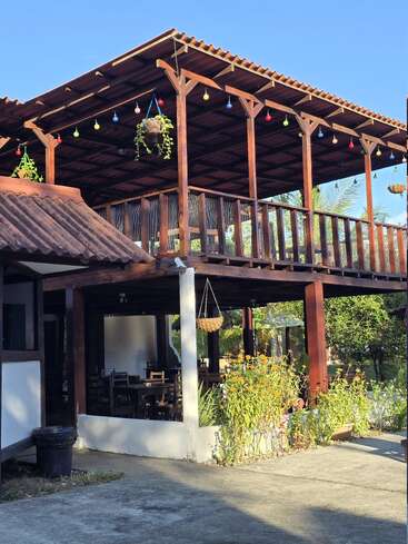 Um terraço de madeira charmoso com telhado de telhas, luzes coloridas penduradas, plantas exuberantes e mesas ao ar livre. A luz do sol ilumina o ambiente aconchegante e convidativo sob um céu azul claro.