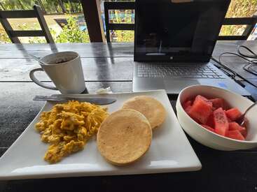Uma cena de café da manhã com ovos mexidos, duas panquecas, uma tigela de melancia, uma xícara de café e um laptop aberto em uma mesa ao ar livre.