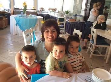 A imagem mostra uma mulher com três filhos sentada em uma mesa de um restaurante, cercada por outras mesas e cadeiras, com uma atmosfera calorosa e convidativa.