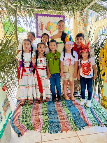 A group of smiling children and an adult, wearing traditional costumes and headpieces, pose together on a colorful rug under palm leaves, celebrating cultural heritage.