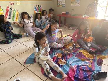A group of children in pajamas sit on blankets and the floor, enjoying snacks and drinks in a classroom decorated with colorful educational posters and crayons.
