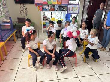 A group of young children in school uniforms sit on colorful chairs in a classroom, some with creative hairstyles and balloons, watched by two adults nearby.