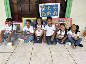 Six children sit on a tiled floor, smiling, in front of colorful classroom posters displaying pictures and drawings. They seem happy and ready for a school activity.