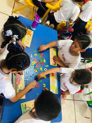 Five young children are gathered around a blue table, playing with colorful alphabet letters and puzzle boards in a lively classroom setting, engaging in learning activities.