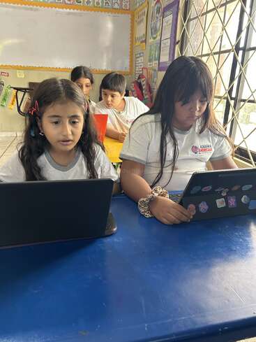 Four young students sit at tables in a colorful classroom, focused on their laptops. They wear white uniforms and appear to be engaged in learning activities.