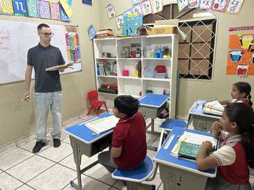 Eine Lehrerin steht an der Tafel und unterrichtet drei aufmerksame Schülerinnen und Schüler, die an blauen Tischen in einem farbenfrohen Klassenzimmer sitzen, das mit Lernplakaten und Materialien gefüllt ist.