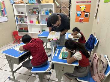 In diesem Klassenzimmer hilft ein Lehrer drei jungen Schülern, die an blauen Schreibtischen arbeiten. Bücher, Rucksäcke und Lernplakate sorgen für eine farbenfrohe, organisierte und ansprechende Lernumgebung.