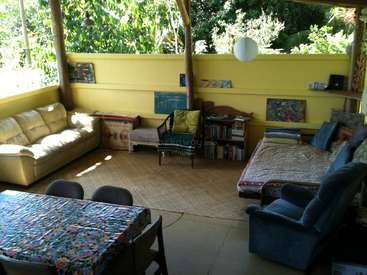 La imagen muestra una acogedora sala de estar al aire libre con una pared amarilla, que incluye un sofá, un sofá cama, una mesa y sillas, rodeados de exuberante vegetación y luz natural.