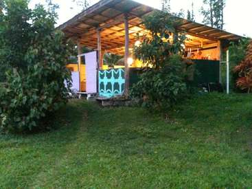 La imagen muestra un pequeño edificio abierto con tejado inclinado, situado en una zona de césped rodeada de árboles y arbustos, con una puerta y una fuente de luz visibles.