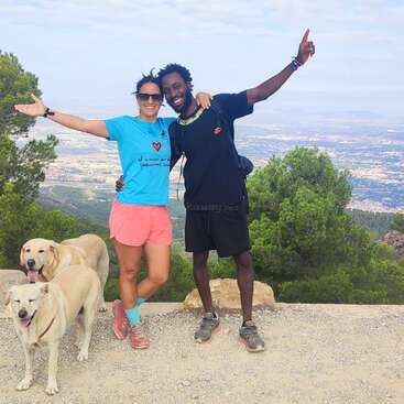 The image depicts a joyful couple standing on a mountain top, with their arms around each other, surrounded by two dogs and a breathtaking view of the city below.