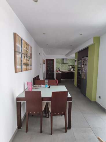 The image depicts a modern kitchen and dining area, featuring a white table with brown chairs, a painting on the wall, and a kitchen with dark cabinets and green accents.