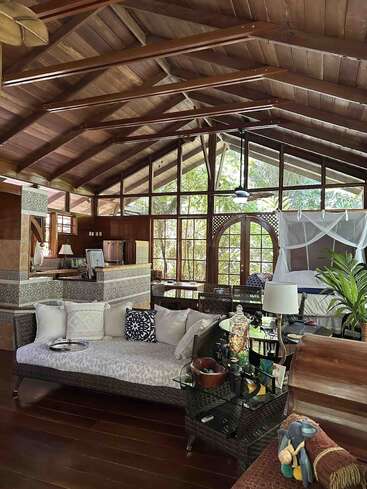 This spacious, cozy cabin features wooden vaulted ceilings, large windows with forest views, wicker furniture, a comfy daybed, canopy bed, kitchen, and inviting, nature-inspired decor.