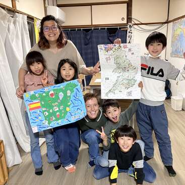 A group of smiling children and two adults pose indoors, holding colorful handmade posters of Spain and England, suggesting a fun educational or cultural classroom activity.
