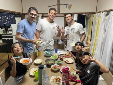 Three adults and three children are smiling around a dining table, enjoying a meal together. The kids are making peace signs. The atmosphere feels friendly and joyful.