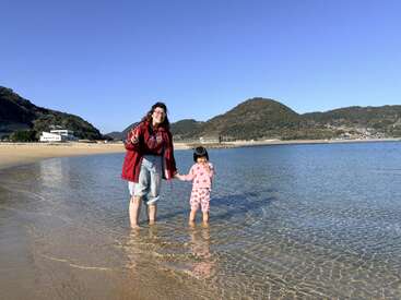 A woman and a young girl stand in shallow water at a beach, smiling under a clear blue sky, with hills and buildings in the background.