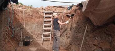 Una mujer está excavando en una profunda zanja arqueológica con un pico. Se ven escaleras, herramientas y un cubo, lo que indica que se están llevando a cabo trabajos de excavación e investigación.
