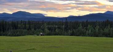 Ein Traktor mäht ein weites grünes Feld unter einem dramatischen Sonnenuntergangshimmel. Der dichte Wald und die fernen Berge bieten eine atemberaubende, ruhige Naturkulisse. Friedliche ländliche Szene.