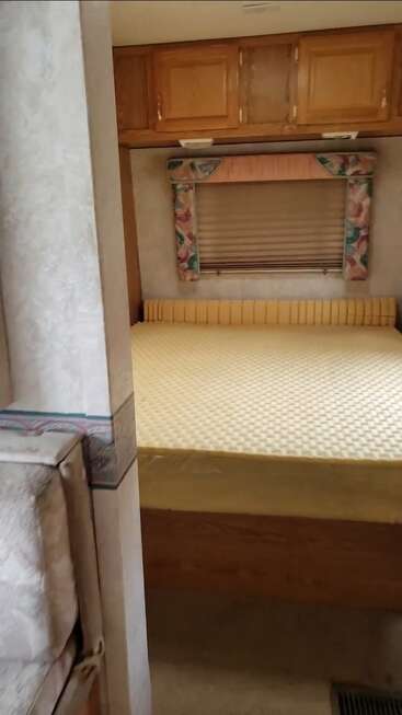 Essa imagem mostra um quarto aconchegante de um trailer com um colchão grande de espuma, armários de madeira em cima, uma janela com cortinas florais e paredes e decoração em tons neutros e suaves.
