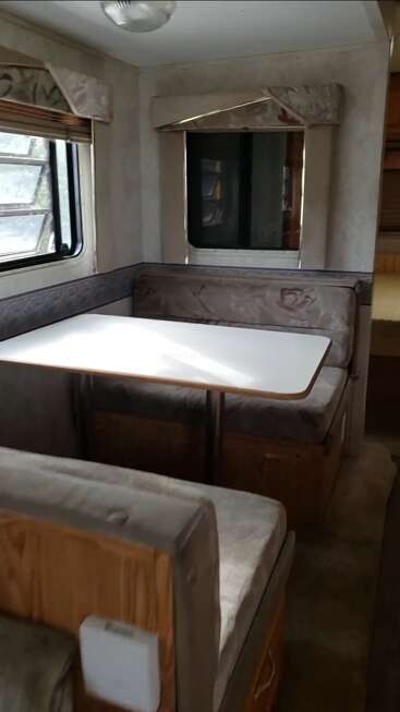 Essa imagem mostra uma área de jantar de um trailer com uma mesa retangular branca, bancos almofadados, duas janelas, cortinas bege e detalhes em madeira, criando um espaço aconchegante.