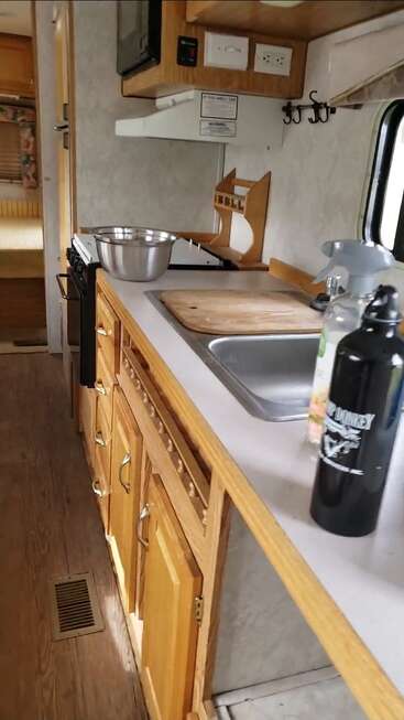 Essa imagem mostra uma cozinha compacta de trailer com armários de madeira, pia, tábua de cortar, eletrodomésticos, tigela de metal, itens de bancada, fogão e um quarto aconchegante na parte de trás.
