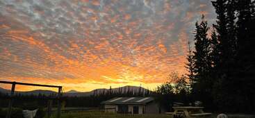 A imagem mostra um pôr do sol deslumbrante com nuvens alaranjadas vibrantes, pinheiros recortados contra o céu, montanhas, um pequeno prédio e mesas de piquenique, criando uma cena ao ar livre serena e tranquila.