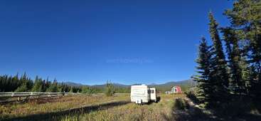 Um trailer branco está parado em um campo aberto e gramado, sob um vasto céu azul, cercado por pinheiros, montanhas, uma cerca de madeira e um galpão vermelho.