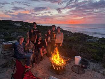 A group of friends gathers around a campfire on a sandy beach at sunset, enjoying each other’s company with the ocean and colorful sky behind them.