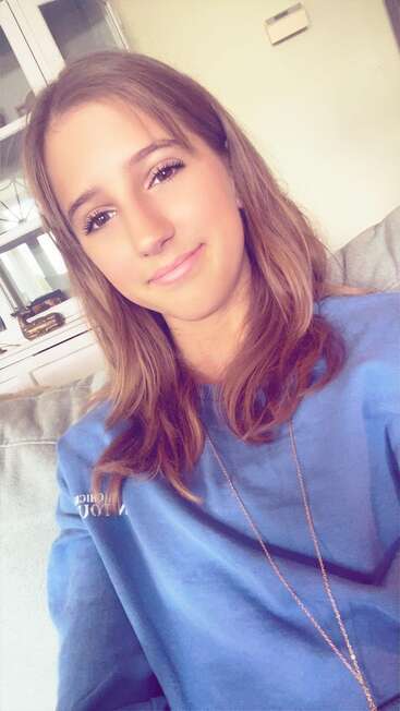 Eine junge Frau mit hellbraunen Haaren sitzt im Haus auf einer Couch, trägt einen blauen Pullover und eine Halskette und lächelt sanft in die Kamera bei natürlichem Licht.